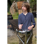 A boy in a striped shirt plays a metal drum outdoors, with a campfire and wooden planks visible in the background.