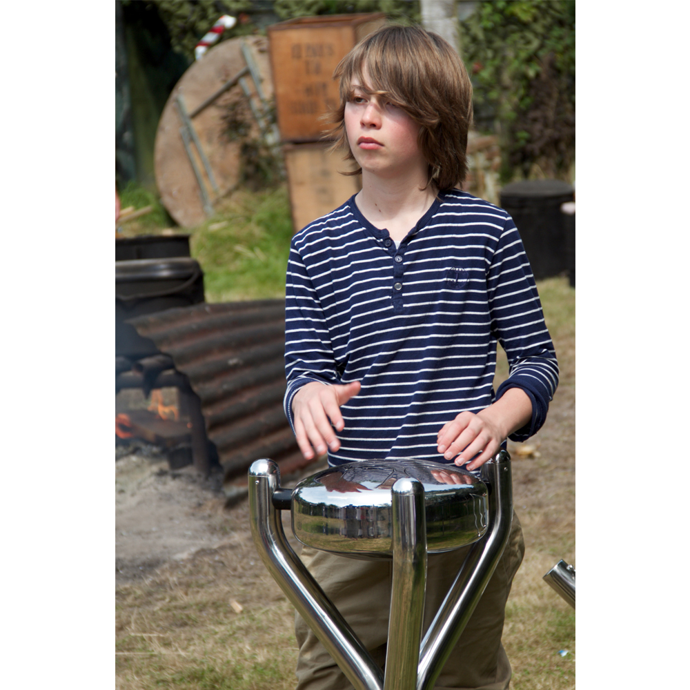 A boy in a striped shirt plays a metal drum outdoors, with a campfire and wooden planks visible in the background.