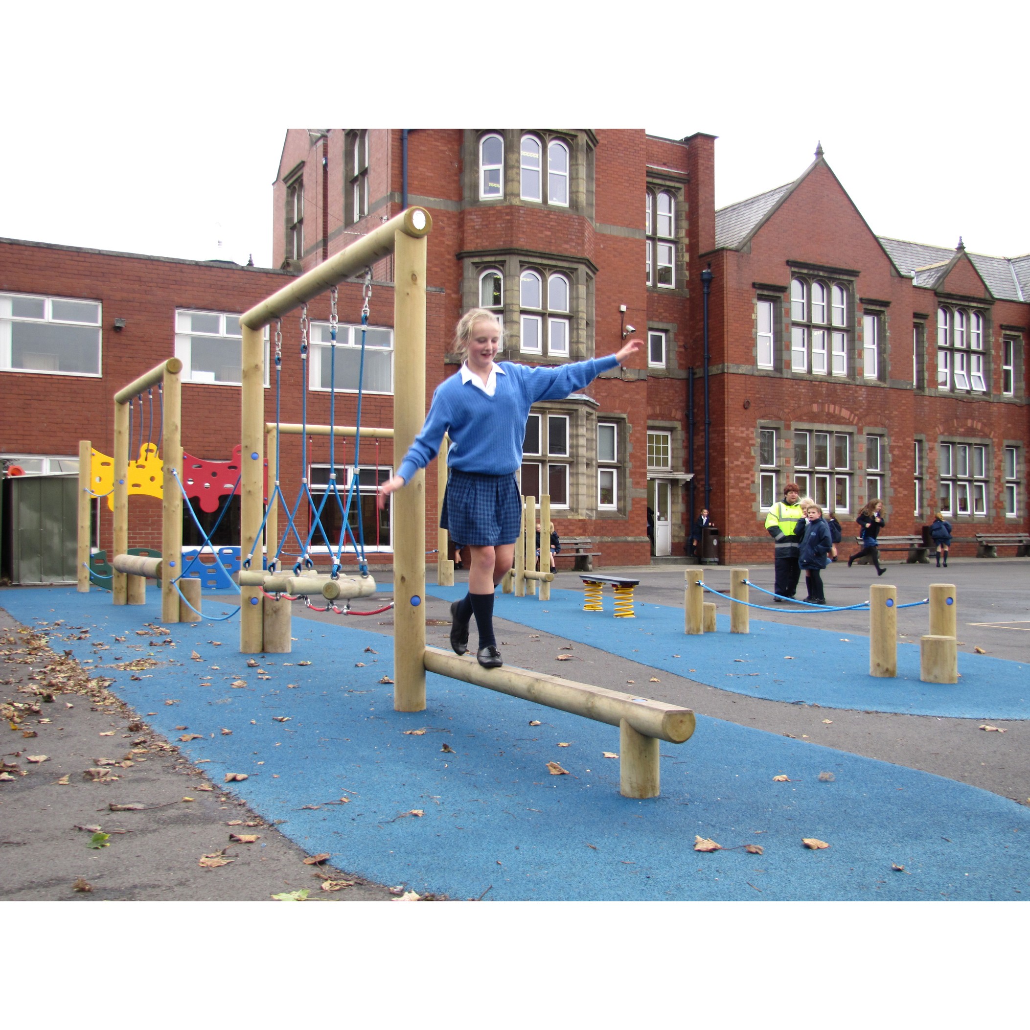 A girl in school uniform balances on a wooden beam in a playground outside a brick school building, with other people and playground equipment in the background.