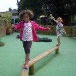 Two young girls balance on a wooden beam in a playground with a green rubberised surface, tyres, and a wooden fence in the background.