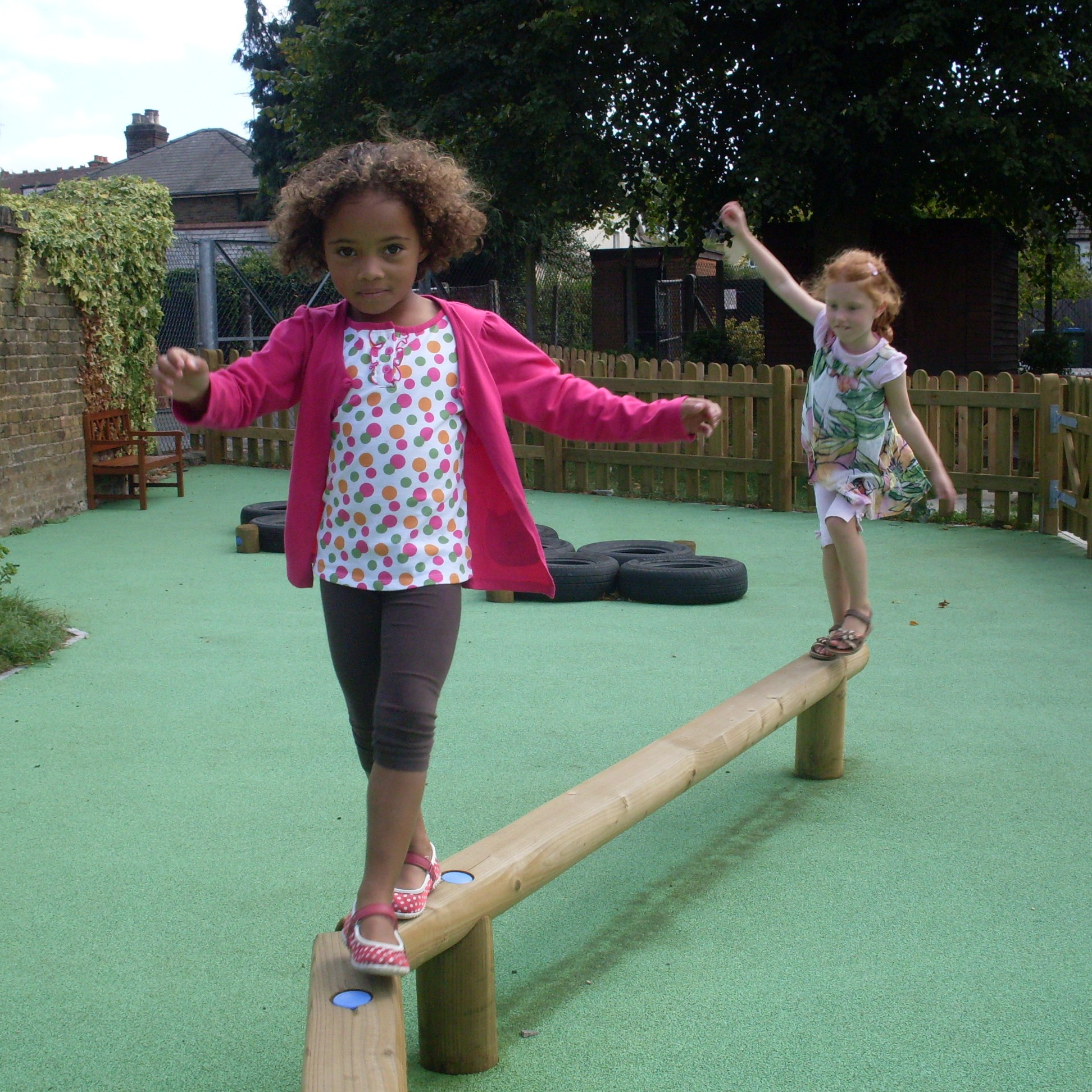 Two young girls balance on a wooden beam in a playground with a green rubberised surface, tyres, and a wooden fence in the background.
