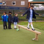 Four children in school uniforms are outside; one girl balances on a wooden beam while three other children wait in a queue behind her.