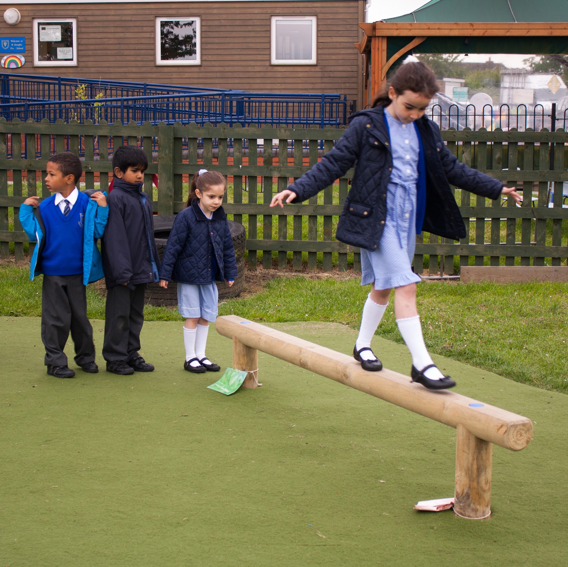 Four children in school uniforms are outside; one girl balances on a wooden beam while three other children wait in a queue behind her.