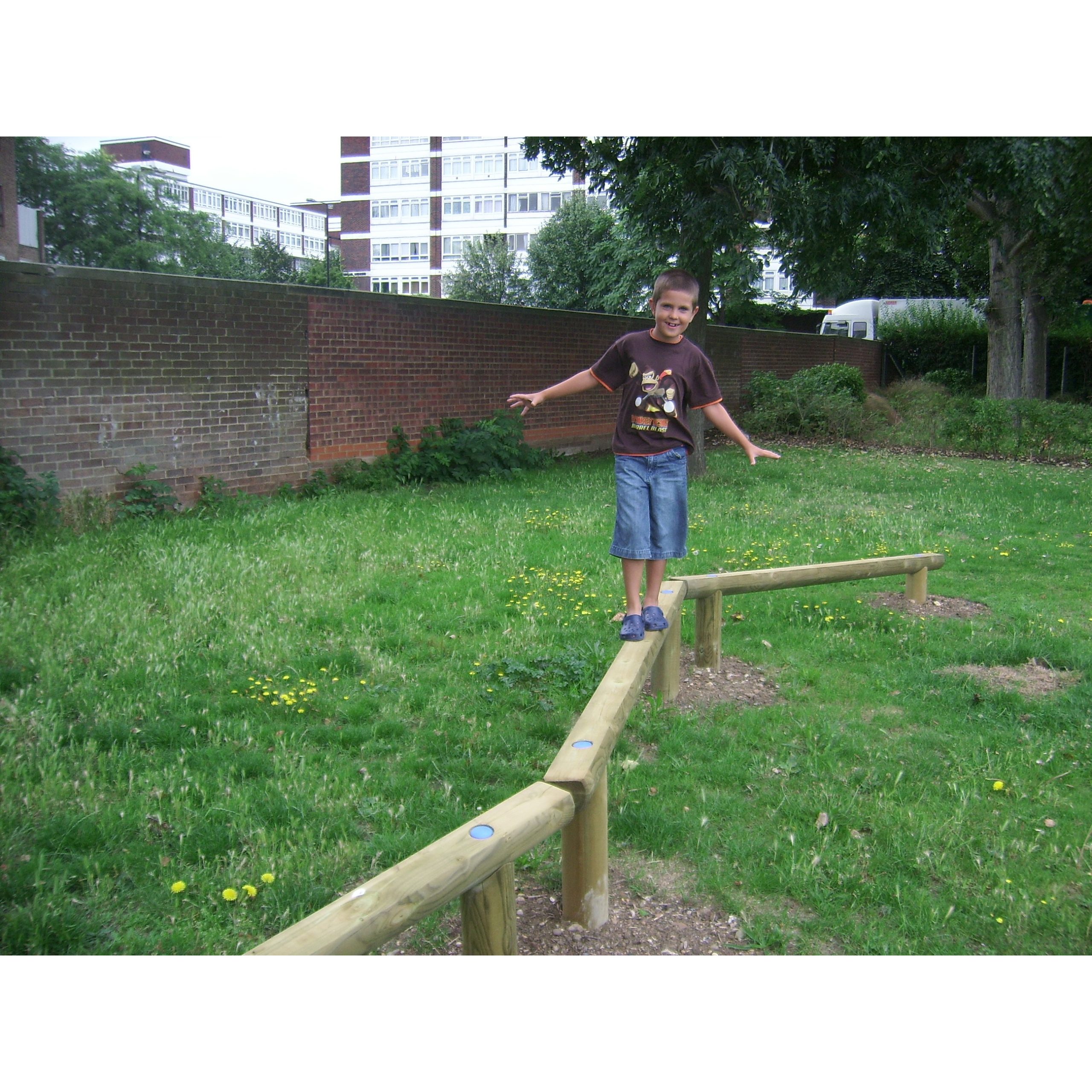 A boy balances on a wooden beam structure in a grassy outdoor area, with buildings and trees in the background.