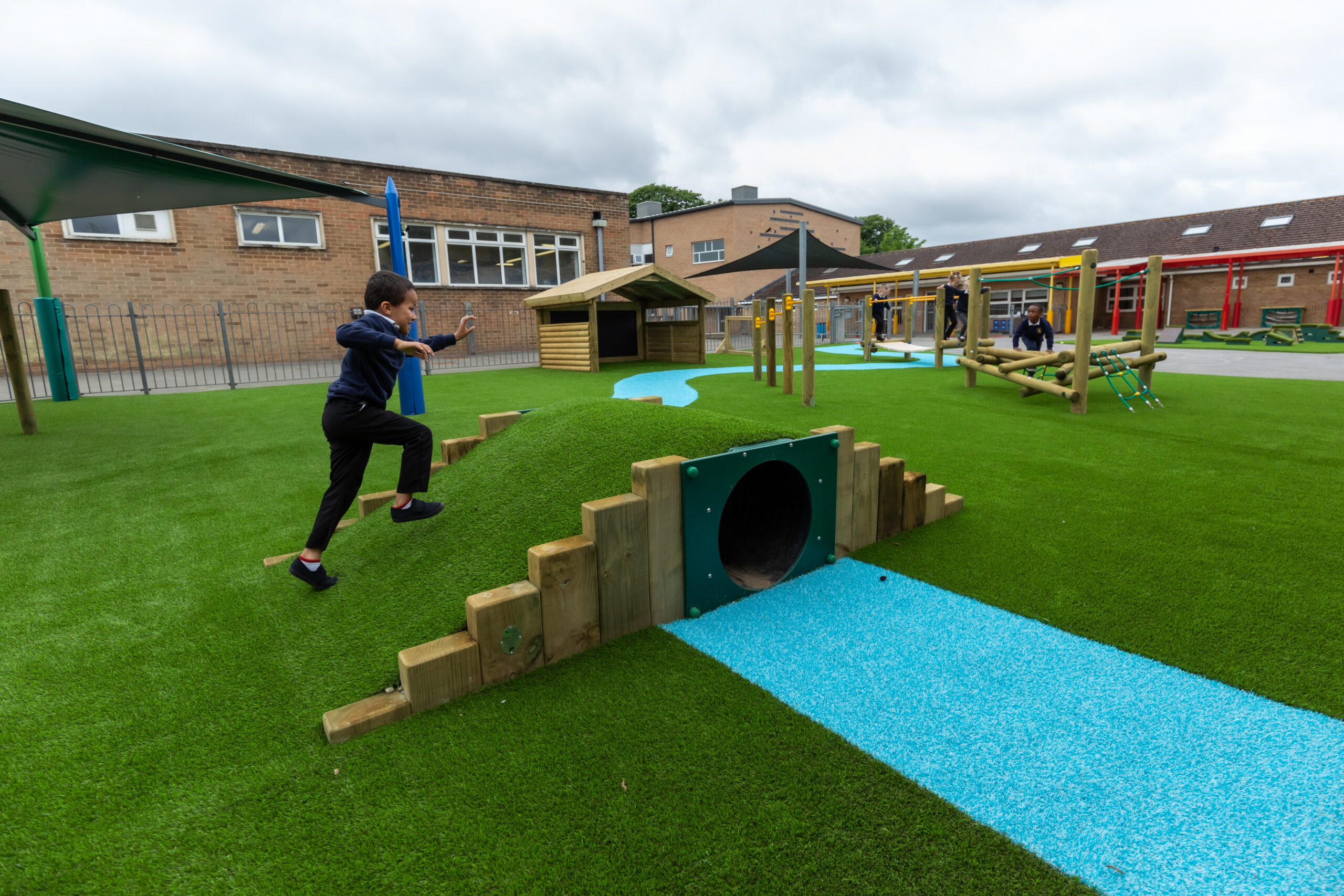 A child runs up Hummock Hill, a small artificial hill with a tunnel, on a modern school playground with green turf and blue paths.