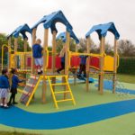 Children in school uniforms play on a colourful playground featuring the Belair Tower 900mm with Poly Slide, plus climbing equipment and slides on a soft rubber surface.