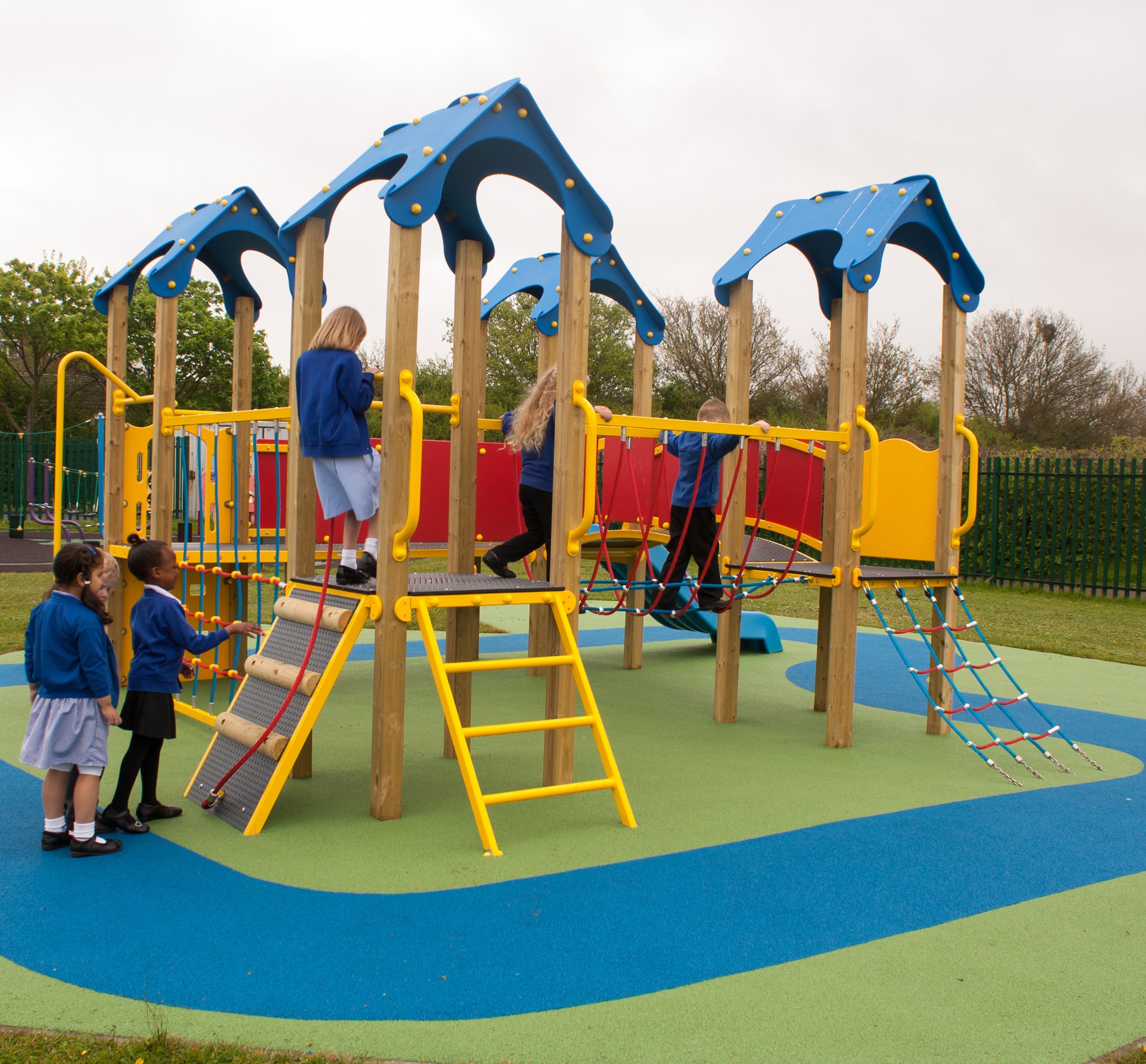 Children in school uniforms play on a colourful playground featuring the Belair Tower 900mm with Poly Slide, plus climbing equipment and slides on a soft rubber surface.