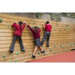 Three children in school uniforms climb a Single Sided Bouldering Wall with green holds in an outdoor play area.