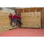 Three children in school uniforms climb horizontally on a Single Sided Bouldering Wall, featuring green holds and a red rubber floor outdoors.