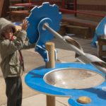 A child in a hooded coat turns a large blue and silver screw pump at an outdoor play area with other play equipment in the background.
