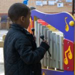 A child wearing a black quilted jacket plays a colourful outdoor xylophone using two mallets.