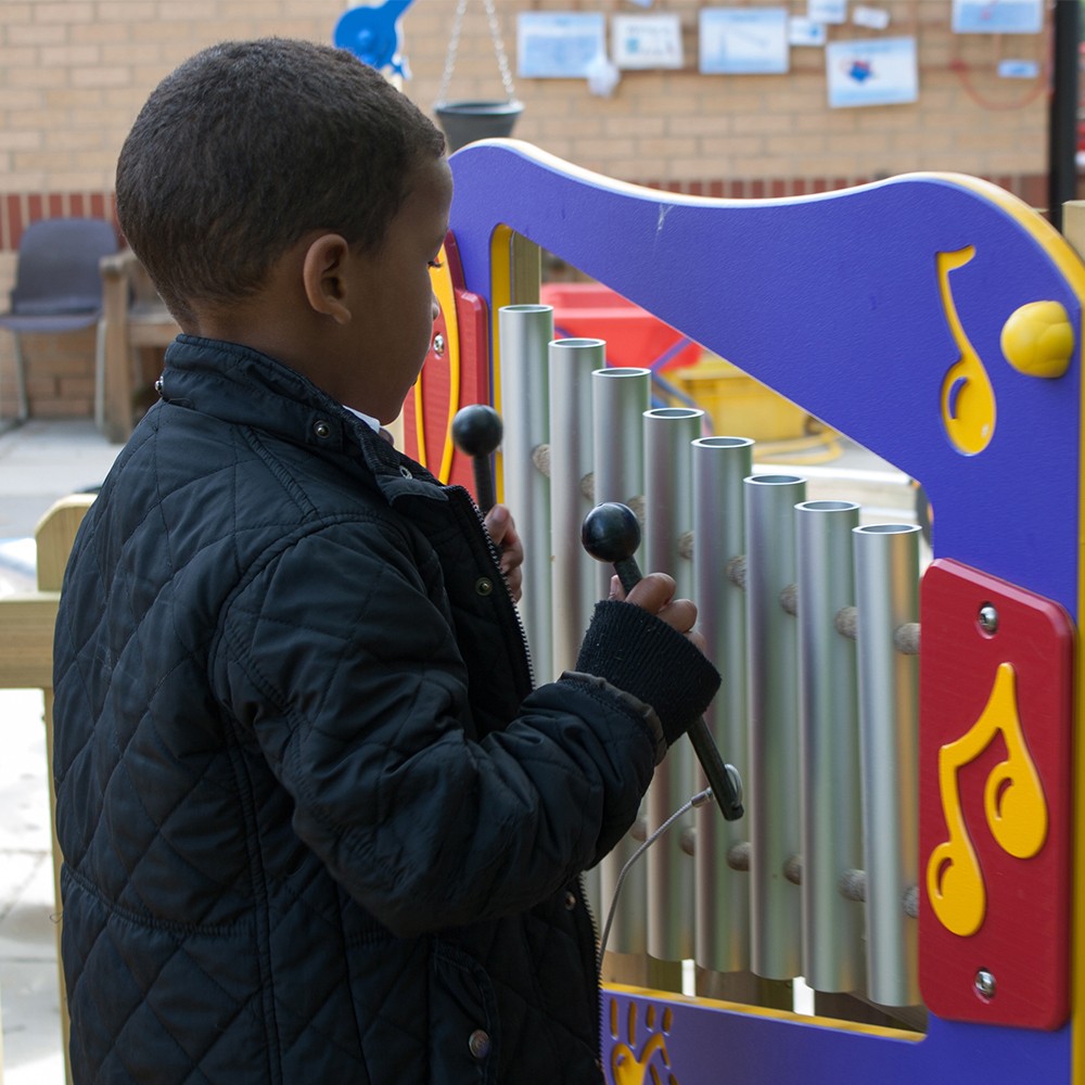A child wearing a black quilted jacket plays a colourful outdoor xylophone using two mallets.