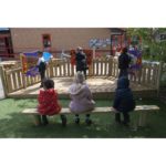 Five children are in an outdoor play area; three sit on a wooden bench, and two stand inside a fenced section with colourful panels.