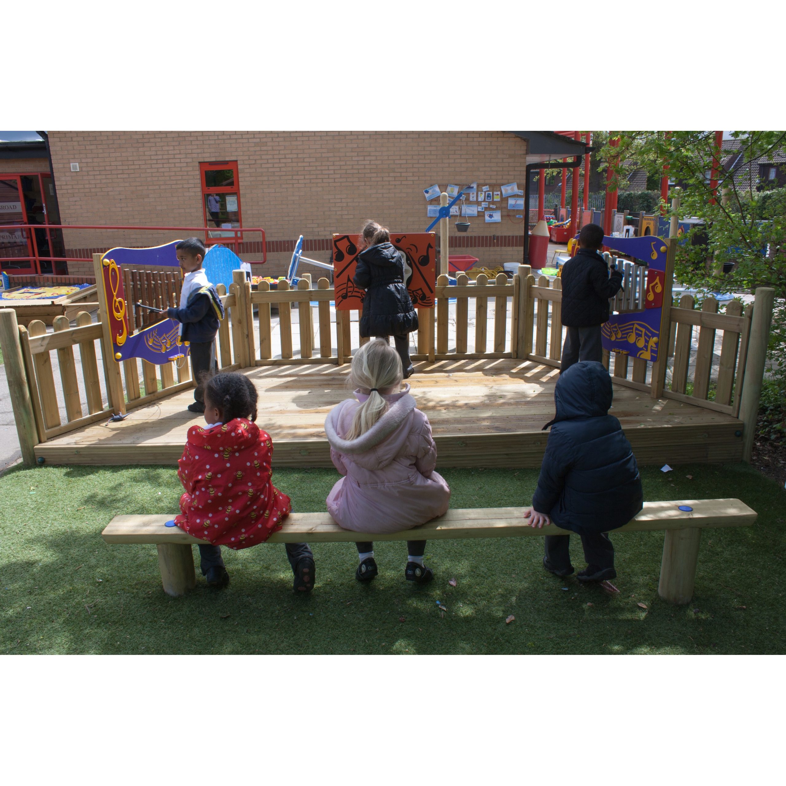 Five children are in an outdoor play area; three sit on a wooden bench, and two stand inside a fenced section with colourful panels.