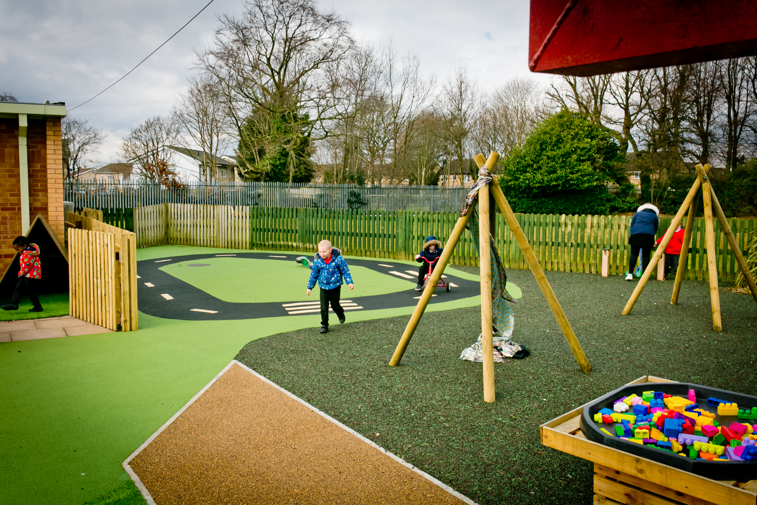 Children play in a fenced outdoor Durabond playground with swings, a toy road, and a box of colourful plastic blocks on a cloudy day.