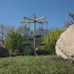 A modern Buccaneer Tower playground climbing structure with ropes and platforms, surrounded by grass, rocks, and trees under a clear blue sky.