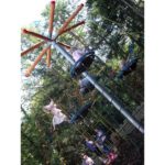 Children climb and play on the tall Buccaneer Tower, a rope and metal playground structure outdoors, surrounded by trees and wood chippings on the ground.