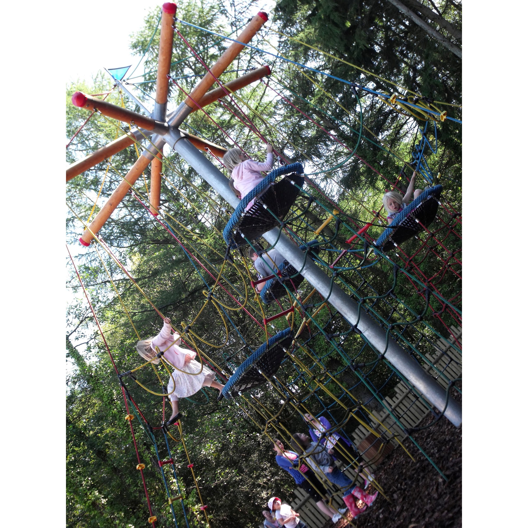Children climb and play on the tall Buccaneer Tower, a rope and metal playground structure outdoors, surrounded by trees and wood chippings on the ground.