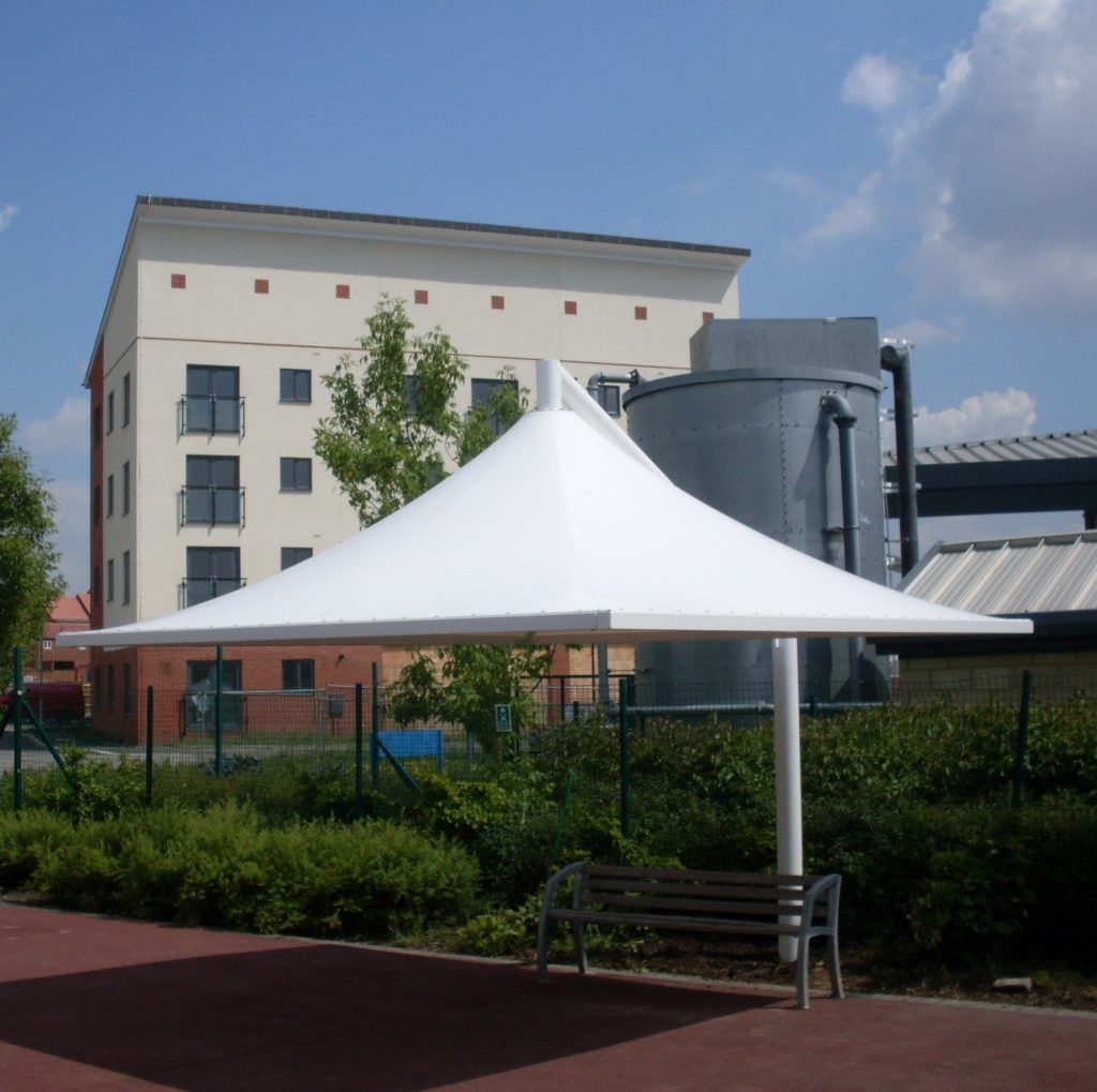 A white, modern canopy structure shades a bench on a paved area, with a building, metal tank, and greenery in the background under a partly cloudy sky.