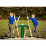 Two young boys in blue jumpers and grey trousers use an outdoor exercise machine together on a grassy field, smiling at the camera.