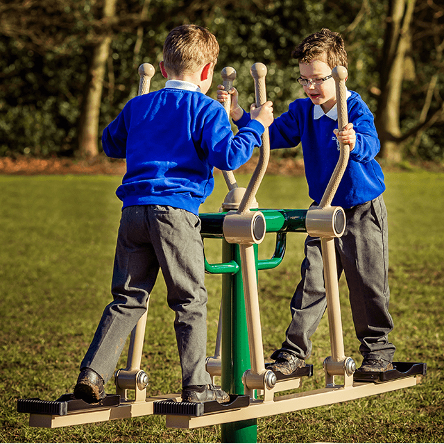 Two young boys in blue jumpers and grey trousers use side-by-side outdoor exercise machines in a grassy park area with trees in the background.