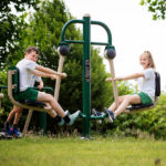 Two children in sports kit use an outdoor exercise machine together in a grassy park area, with greenery in the background.