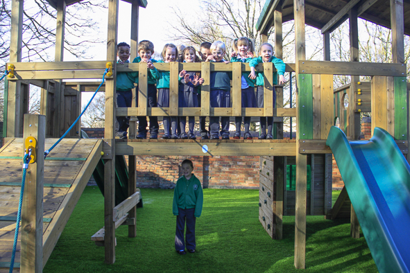A group of children stand on a wooden playground structure, while one Young Explorer stands alone on the artificial grass below.