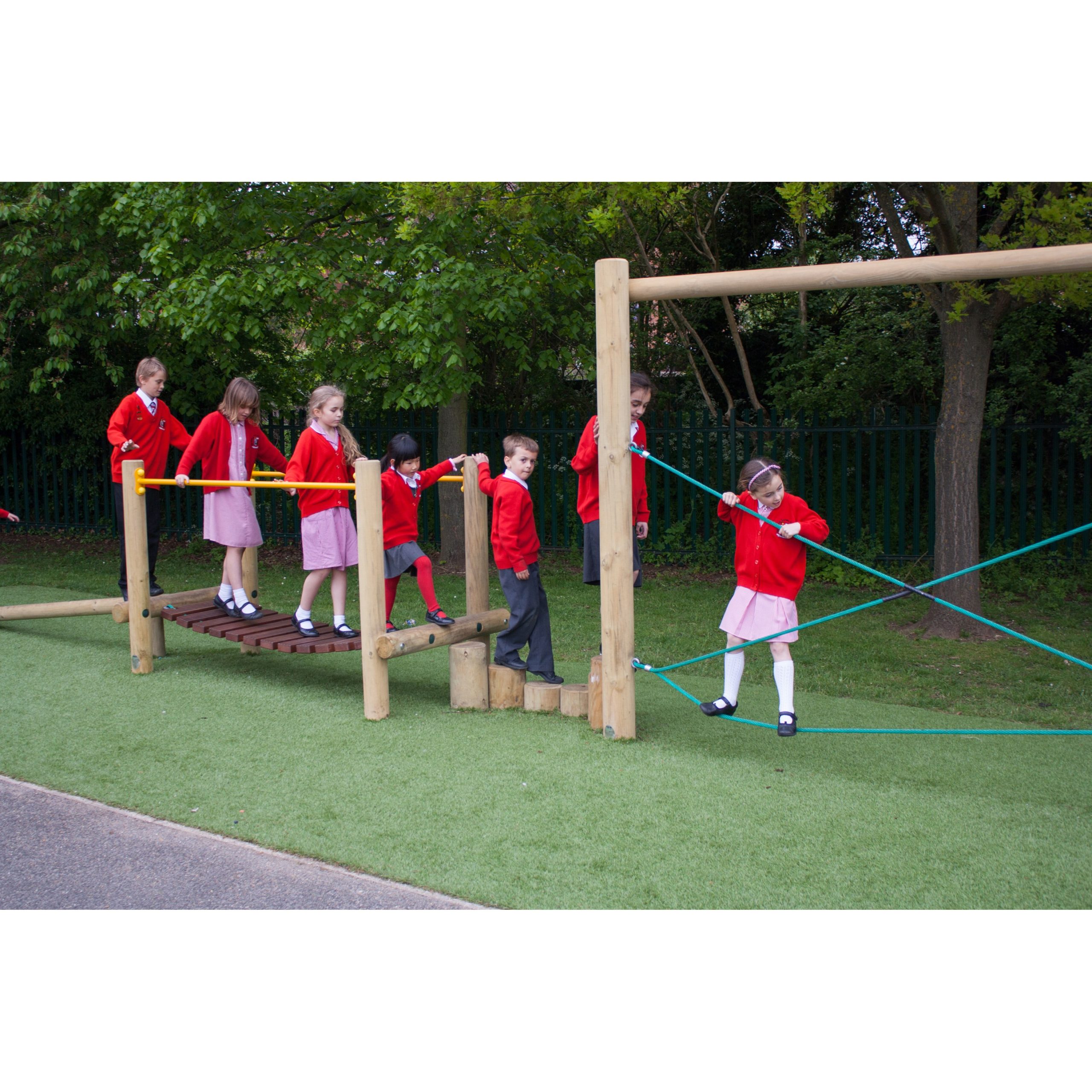 Several children in red school uniforms play on a wooden climbing frame outdoors on artificial grass near trees.
