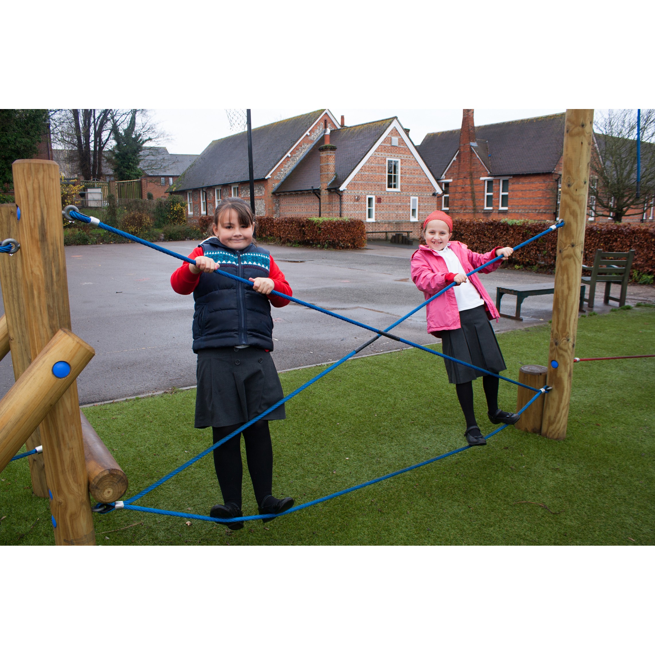 Two school-age girls stand on a rope climbing structure on a playground, holding blue ropes, with school buildings visible in the background.