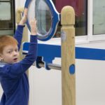 A young boy in a blue jumper reaches up to touch a large circular magnifying glass mounted on a wooden and metal structure indoors.
