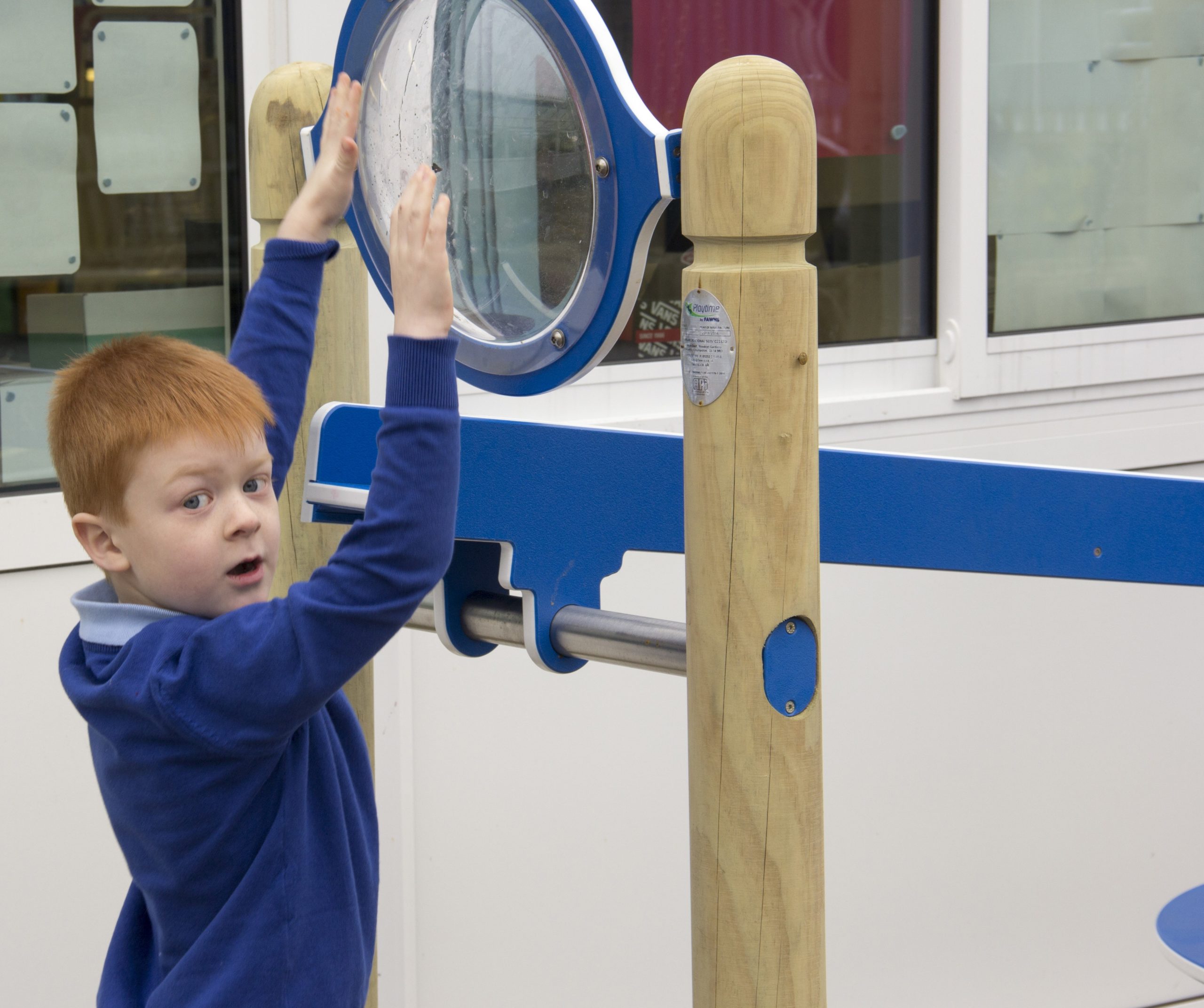 A young boy in a blue jumper reaches up to touch a large circular magnifying glass mounted on a wooden and metal structure indoors.