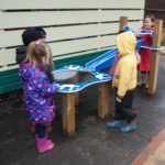 Five young children stand around a blue water play table outdoors, engaging with the structure on a wet playground surface.