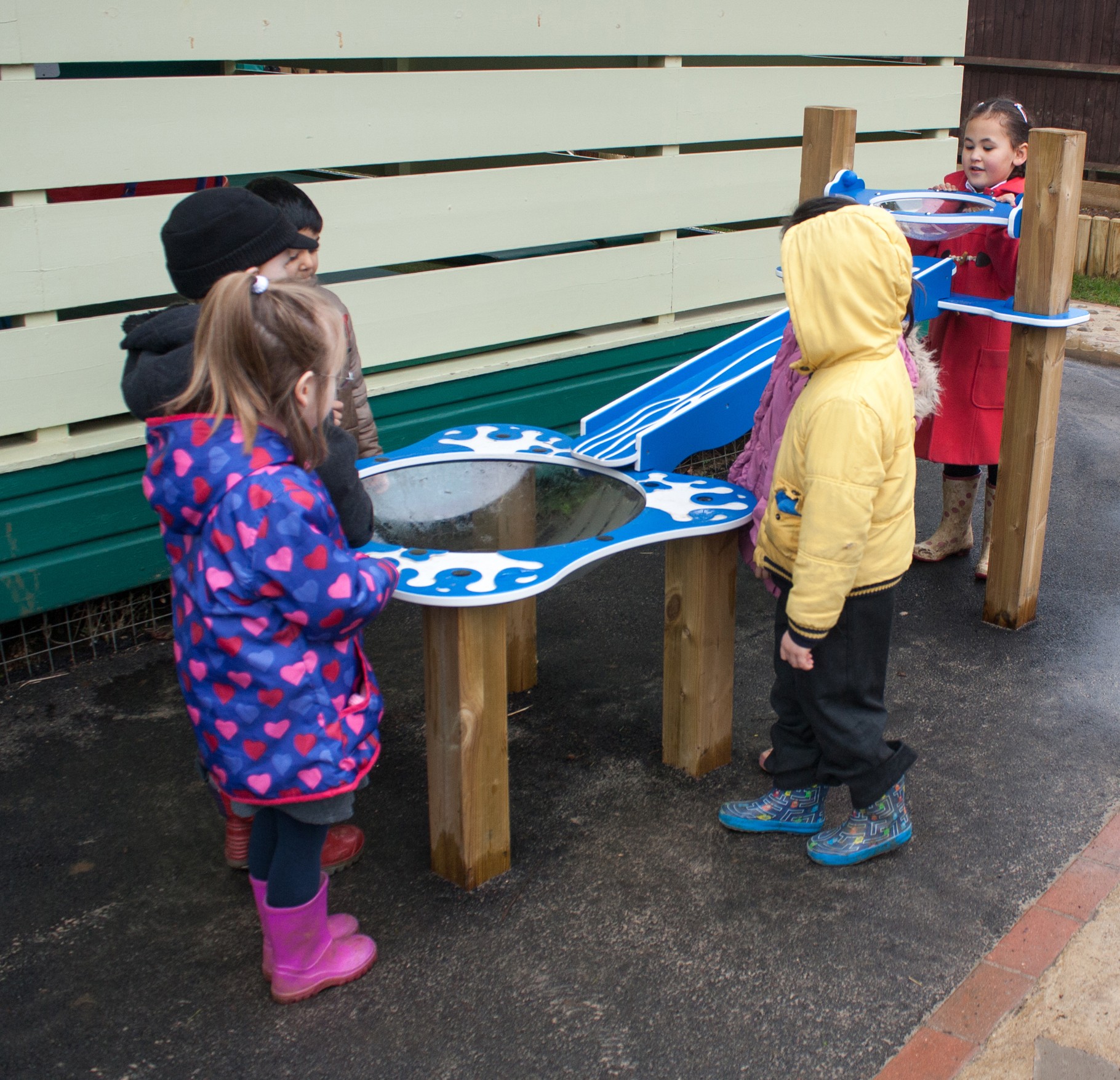 Five young children stand around a blue water play table outdoors, engaging with the structure on a wet playground surface.