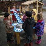 Six children in outdoor clothing stand around a blue and white water play table at a playground, with a wooden shelter in the background.