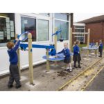 Children in school uniforms play with blue outdoor water play equipment beside a building.