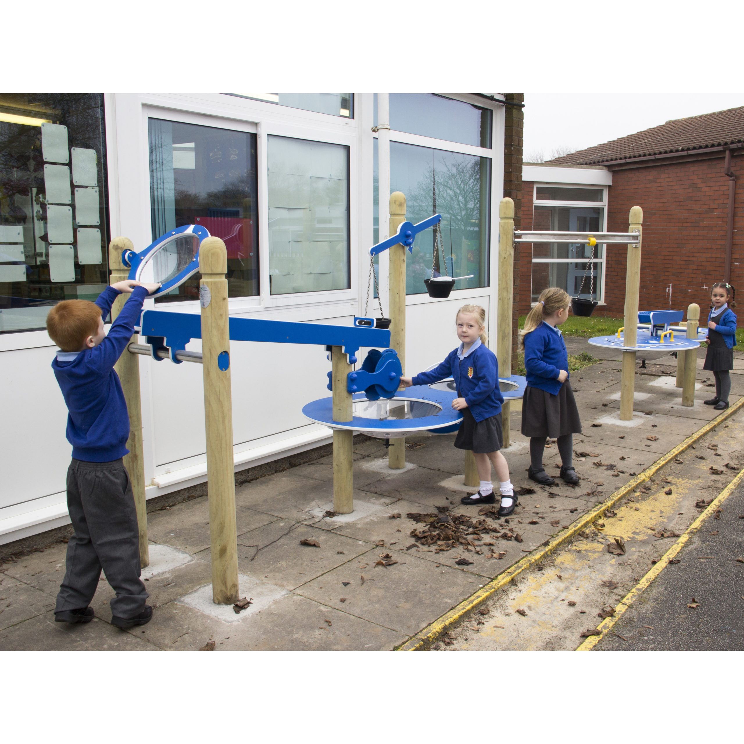 Children in school uniforms play with blue outdoor water play equipment beside a building.