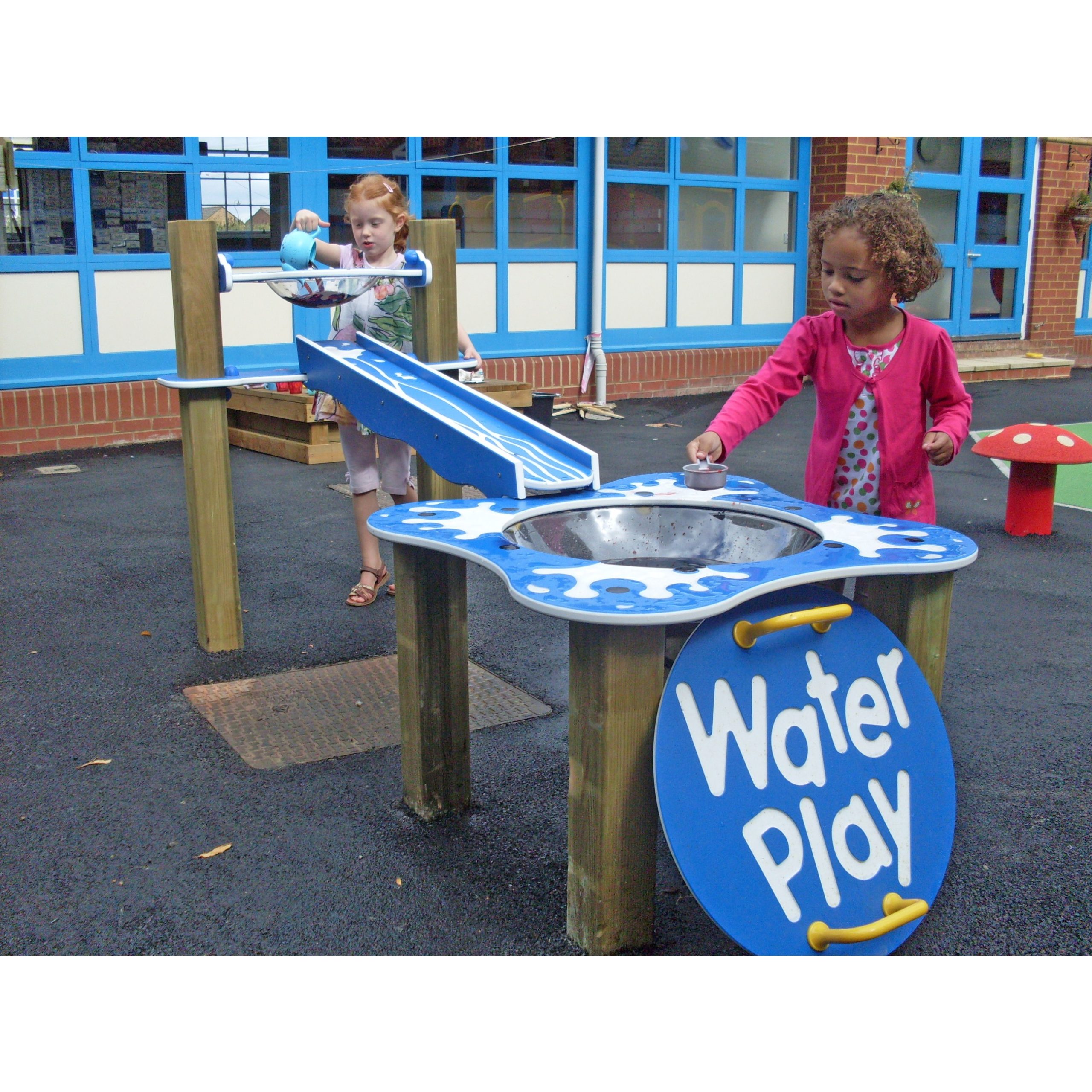 Two young girls play with a blue water play station outdoors, using tools to move water along chutes and into a basin.