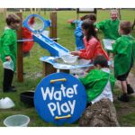 Six young children in colourful smocks play with water at an outdoor water play station, pouring and collecting water using jugs and containers.