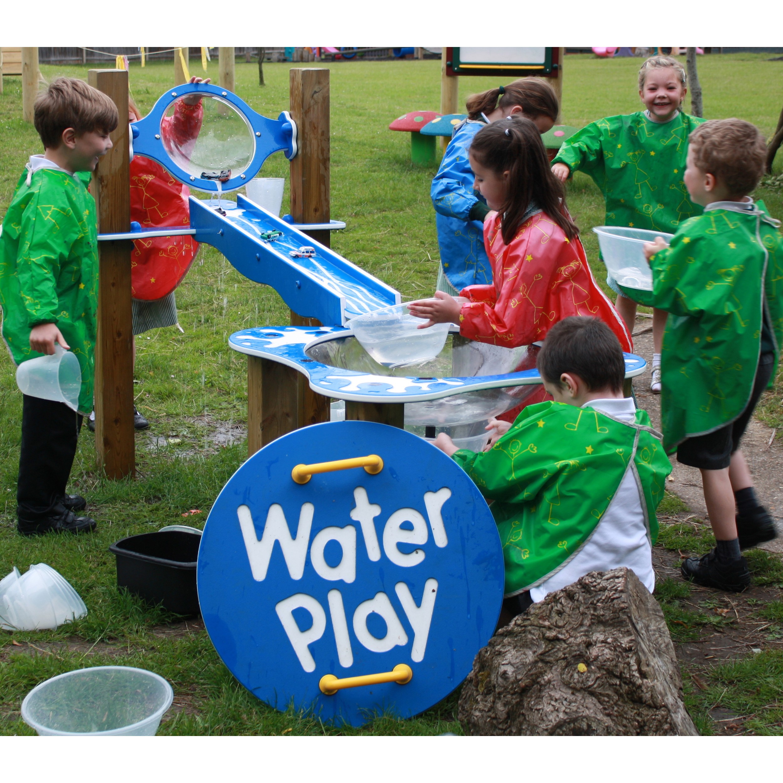 Six young children in colourful smocks play with water at an outdoor water play station, pouring and collecting water using jugs and containers.