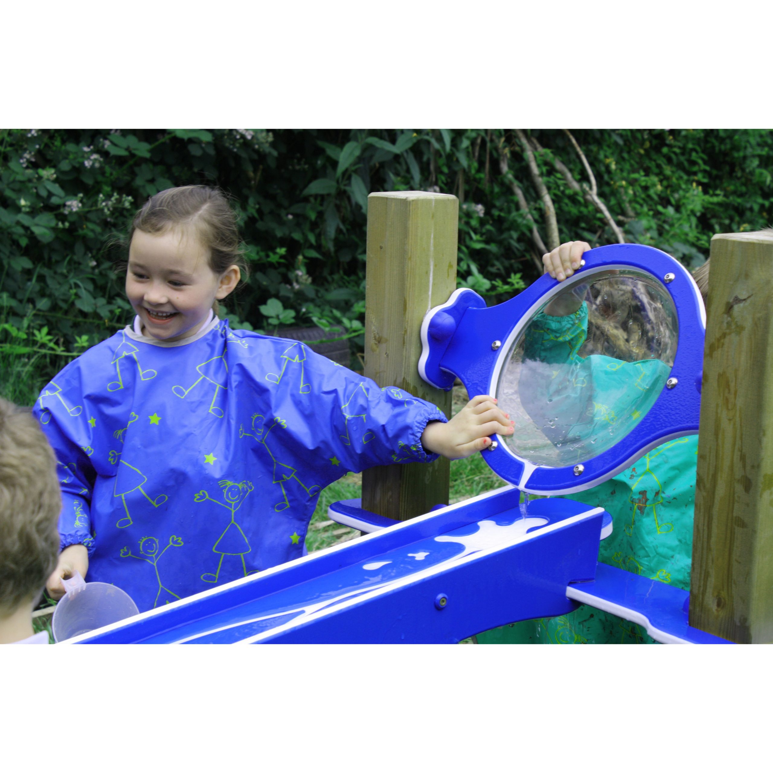 Two children in waterproof overalls play with a blue water activity table outdoors, one smiling while pouring water down a chute, the other partially hidden behind a circular panel.