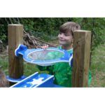 A young boy wearing a green apron stands outdoors, looking through a clear plastic window mounted between two wooden posts.