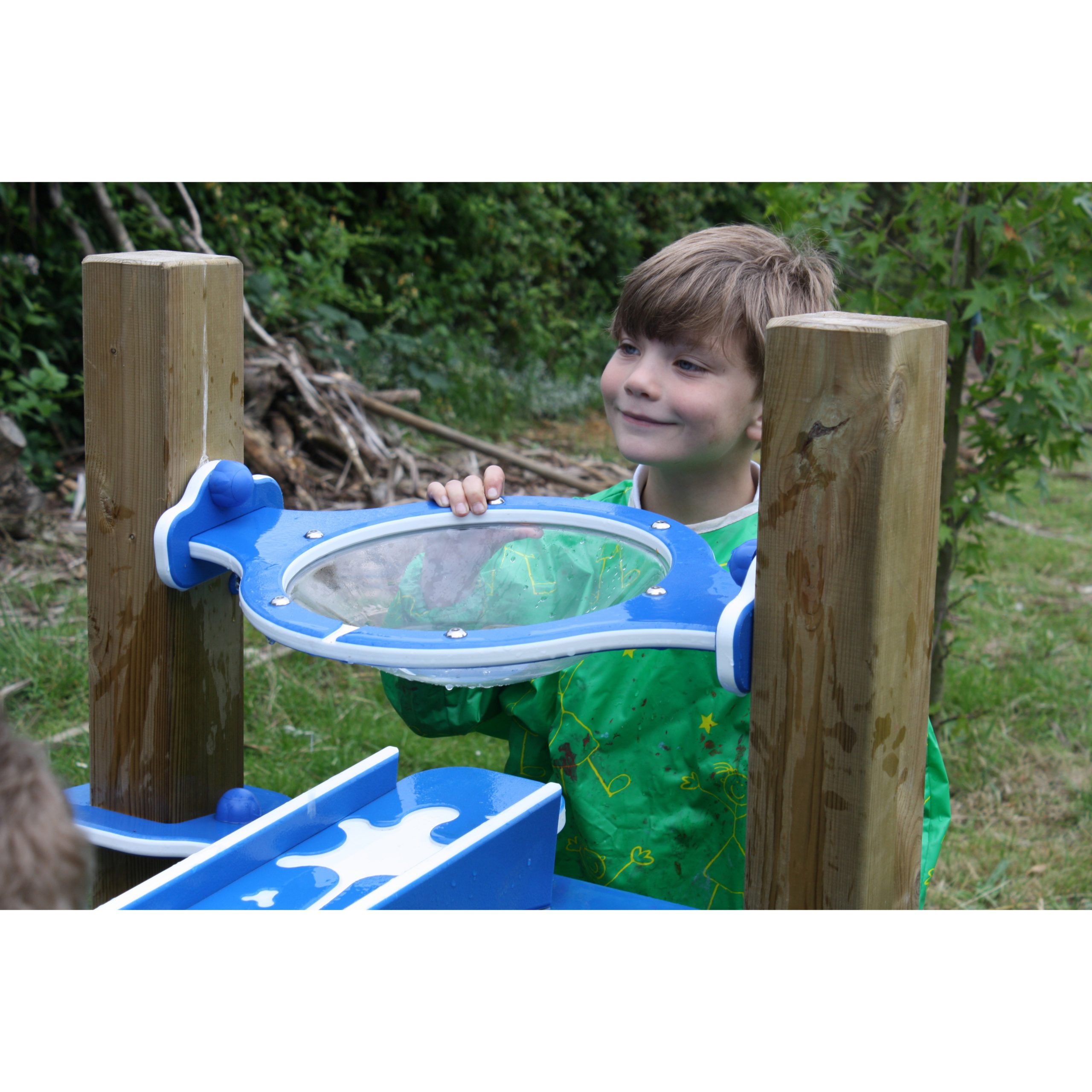 A young boy wearing a green apron stands outdoors, looking through a clear plastic window mounted between two wooden posts.