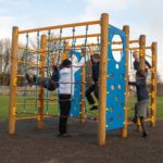 Five children play on a wooden and rope Discovery Space Frame with blue panels in an outdoor playground on a cloudy day.