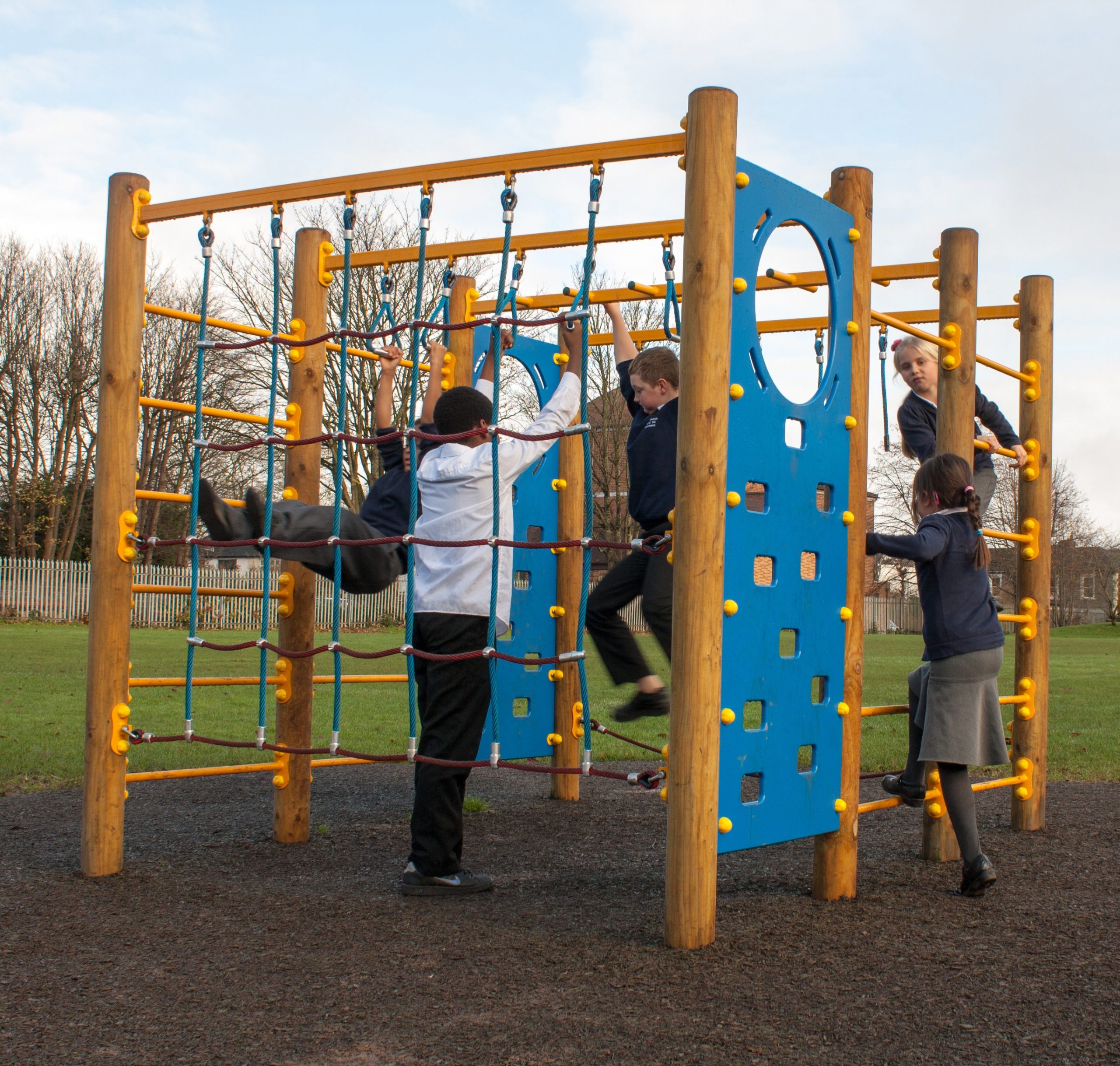Five children play on a wooden and rope Discovery Space Frame with blue panels in an outdoor playground on a cloudy day.