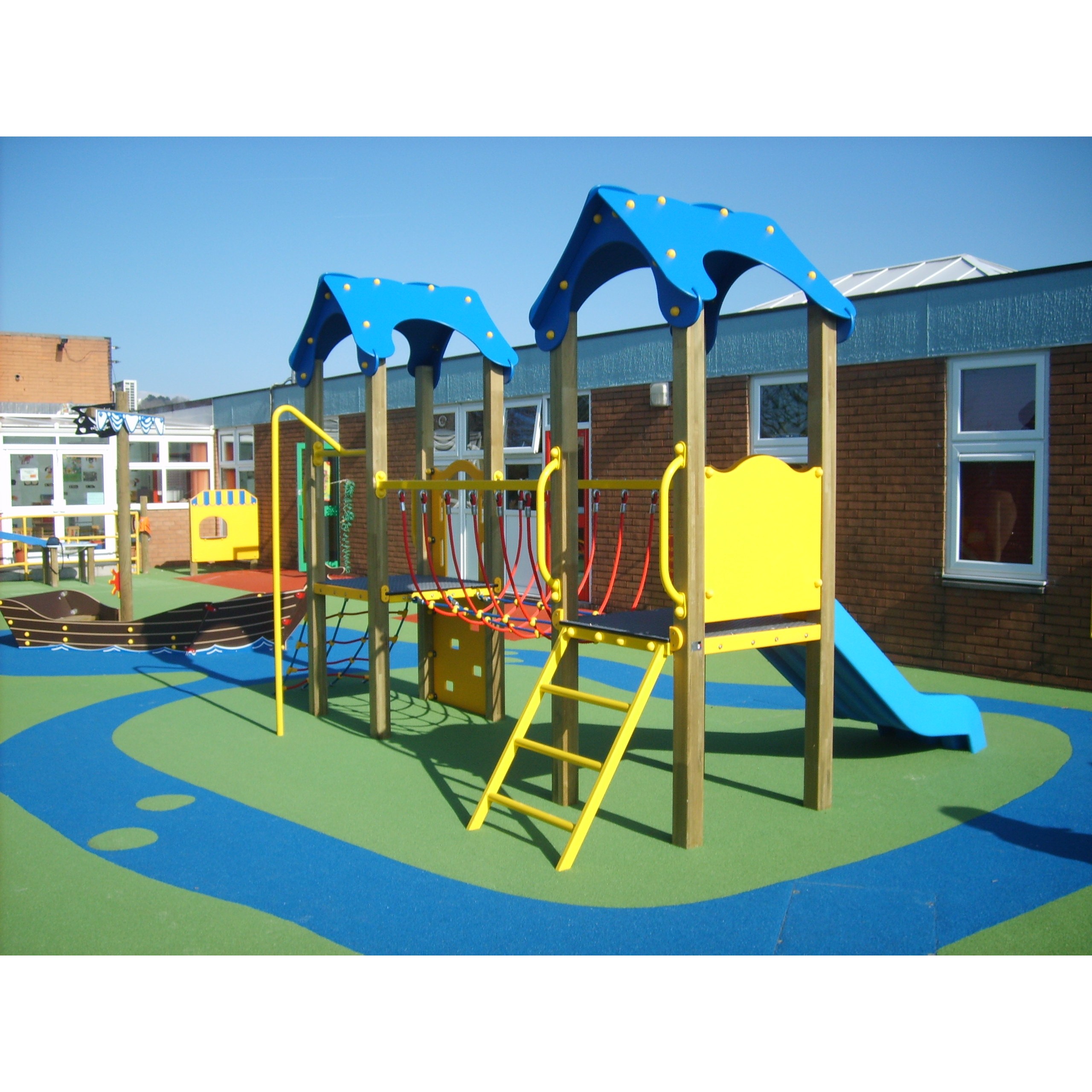 Outdoor playground featuring the Dixie Tower 1200mm with Poly Slide, climbing structures, and blue and yellow roofs on a colourful rubber surface, next to a brick building under a clear sky.