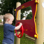 A young child stands outdoors and plays with a red and yellow steering wheel attached to a wooden climbing frame, simulating driving.