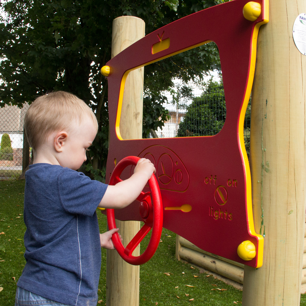 A young child stands outdoors and plays with a red and yellow steering wheel attached to a wooden climbing frame, simulating driving.