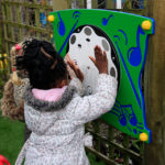 Two children play with an outdoor interactive musical wall panel featuring buttons and musical note designs.