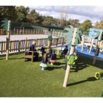 Children in school uniforms sit on mushroom-shaped seats around a teacher outdoors in a fenced playground with artificial grass and play structures.