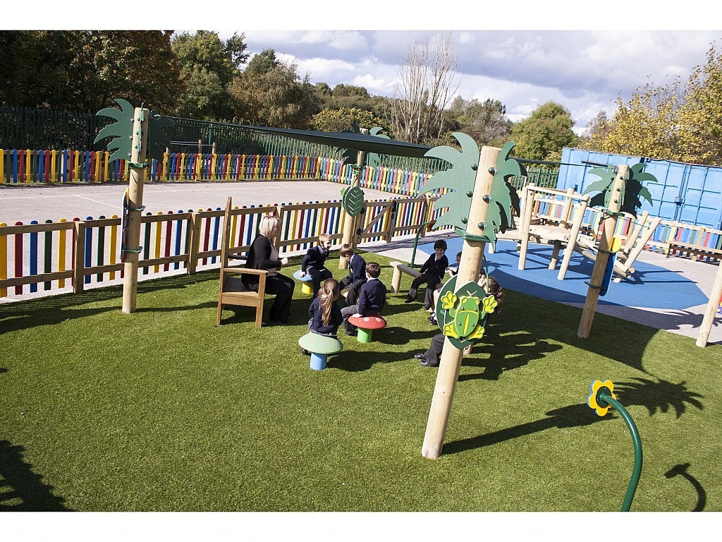 Children in school uniforms sit on mushroom-shaped seats around a teacher outdoors in a fenced playground with artificial grass and play structures.